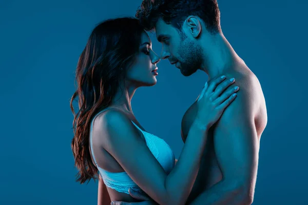 Hemdloser Mann und sexy Mädchen in weißem BH umarmen sich, während sie isoliert von Angesicht zu Angesicht auf dunkelblau stehen — Stockfoto