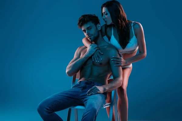 Schöner, hemdloser Mann sitzt auf einem Stuhl, während leidenschaftliches Mädchen in weißen Dessous ihn auf blauem Hintergrund umarmt — Stockfoto