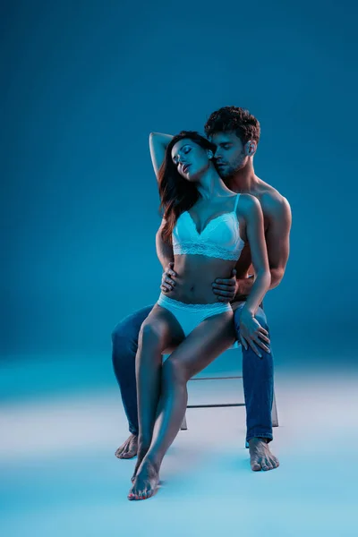 Hombre sin camisa y chica seductora en lencería blanca sentada en la silla y abrazándose sobre fondo azul - foto de stock