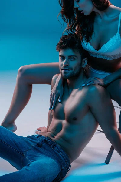 Sexy homem sentado no chão e sorrindo para a câmera enquanto sedutor menina abraçando-o no fundo azul — Fotografia de Stock