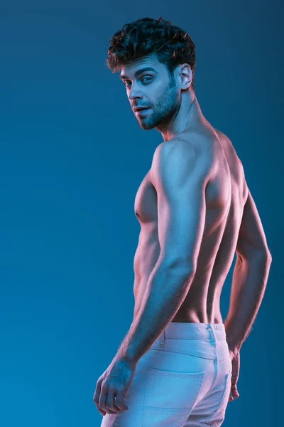 Без сорочки, м'язистий чоловік у білих джинсах дивиться на камеру ізольовано на синьому — стокове фото