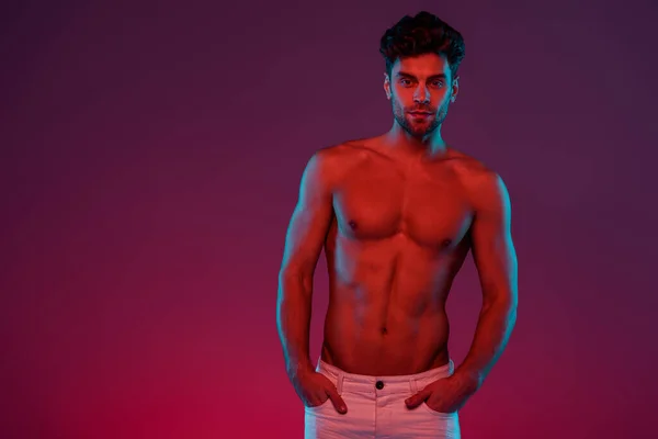 Без сорочки, сексуальний чоловік дивиться на камеру, тримаючи руки в кишенях на фіолетовому фоні — стокове фото