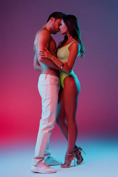 Full length view of shirtless man in white jeans and seductive girl in swimsuit embracing while standing on blue on purple background with gradient — Stock Photo