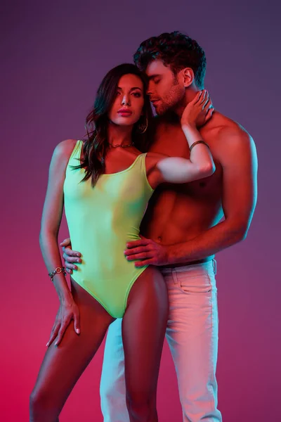 Shirtless man in white jeans hugging seductive girl in swimsuit touching his neck on purple background — Stock Photo