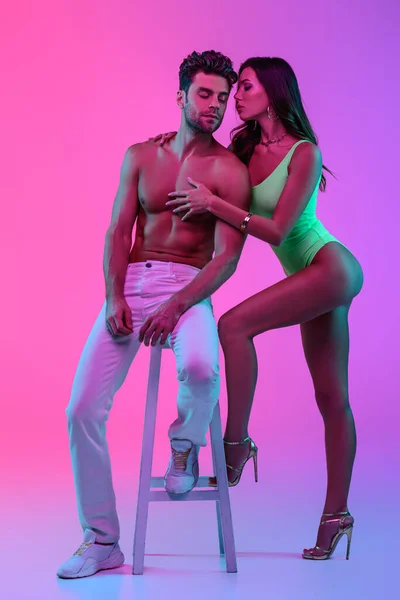 Full length view of sexy girl in green swimsuit hugging shirtless man sitting on bar stool on pink and purple background — Stock Photo