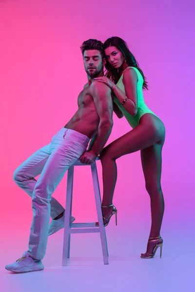 Full length view of passionate girl in green swimsuit touching shoulders of shirtless man sitting on bar stool on pink and purple background — Stock Photo