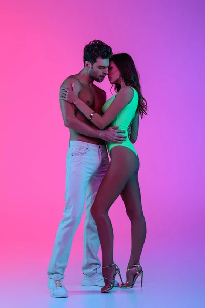 Full length view of sexy shirtless man in white jeans and seductive girl in green swimsuit hugging on pink and purple background — Stock Photo