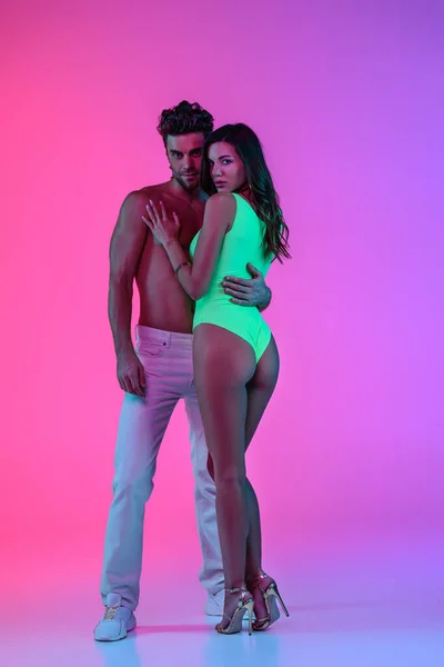 Piena vista lunghezza dell'uomo sexy in jeans bianchi abbracciando seducente ragazza in costume da bagno verde su sfondo rosa e viola — Foto stock