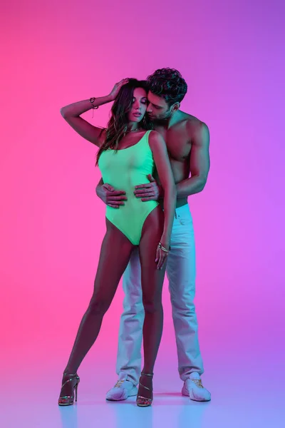 Visão de comprimento completo do homem sem brilho sexy em jeans brancos abraçando menina sedutora em traje de banho verde no fundo rosa e roxo — Fotografia de Stock