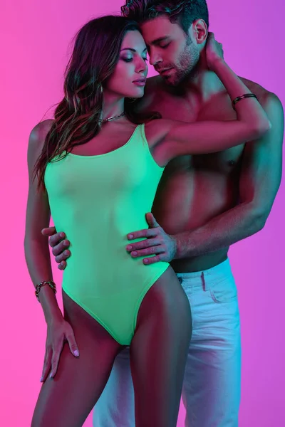 Menina sensual em maiô verde tocando pescoço de homem sem camisa abraçando-a no fundo rosa e roxo — Fotografia de Stock