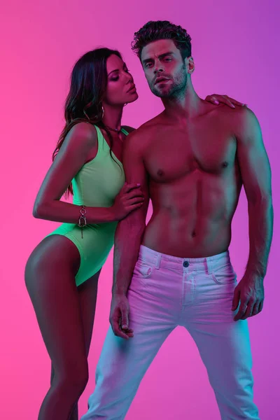 Seductive girl in swimsuit embracing shirtless man in white jeans on pink and purple background — Stock Photo