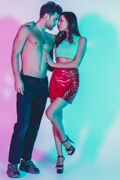 Sexy Mädchen in rotem Minirock und hemdloser Mann in dunkelblauen Jeans, die sich auf dem Hintergrund mit türkisfarbenen und violetten Schatten umarmen — Stockfoto
