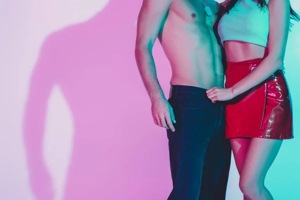 Cropped view of sexy girl in red mini skirt touching jeans of shirtless man on background with violet shadow — Stock Photo