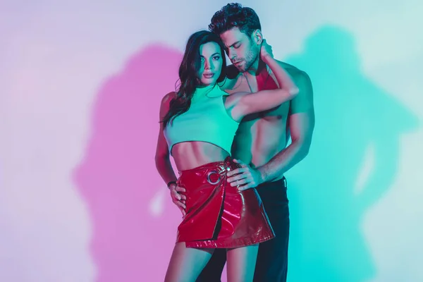 Shirtless man touching hips of sexy girl embracing his neck and looking at camera on blue background with turquoise and violet shadows — Stock Photo