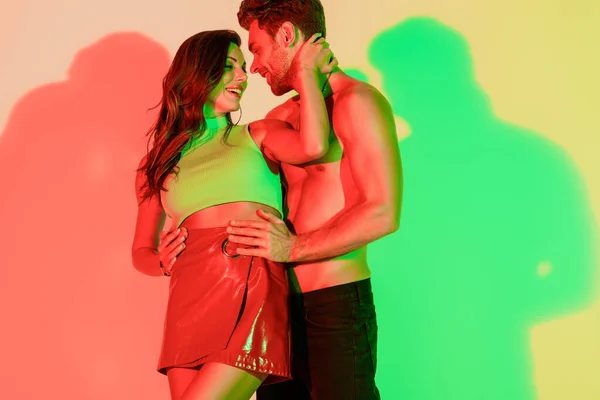 Sexy, homem sem camisa tocando quadris de elegante, menina sexy abraçando seu pescoço no fundo amarelo com sombras vermelhas e verdes — Fotografia de Stock