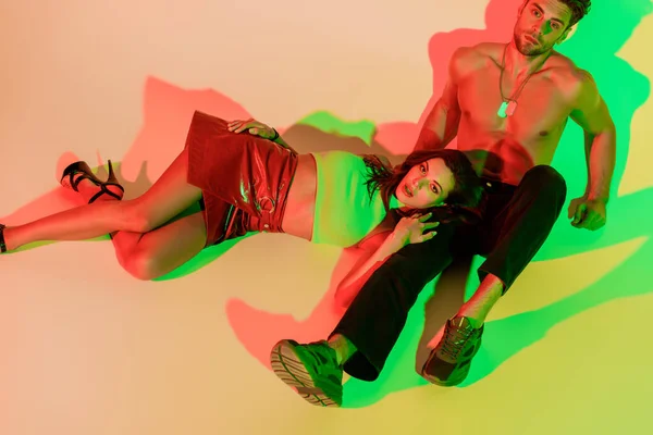 Menina bonita e elegante olhando para a câmera enquanto deitado em sexy, homem sem camisa sentado no fundo amarelo com sombras vermelhas e verdes — Fotografia de Stock