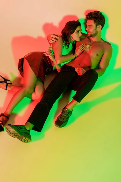 Sexy, trendy girl and hadsome, shirtless man looking at each other while lying on yellow background with red and green shadows — Stock Photo