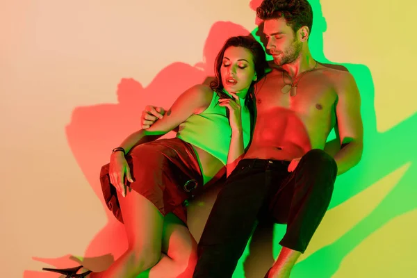 Sexy man embracing sensual, stylish girl while lying on yellow background with red and green shadows — Stock Photo