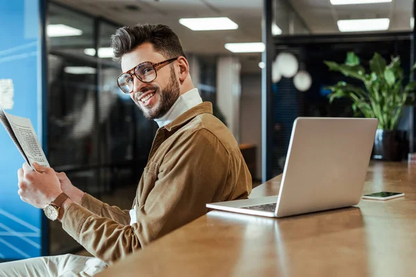 IT worker with newspaper smiling and looking at camera at table with laptop in coworking space — Stock Photo