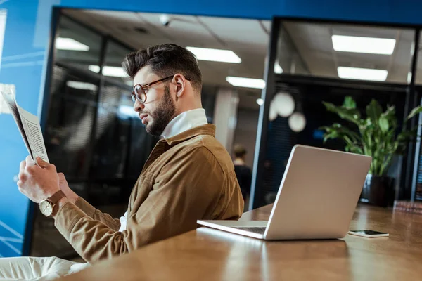 Trabajador de TI leyendo periódico en la mesa con portátil en el espacio de coworking - foto de stock