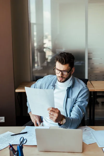 Concentrated IT worker holding papers near laptop at table in coworking space — Stock Photo