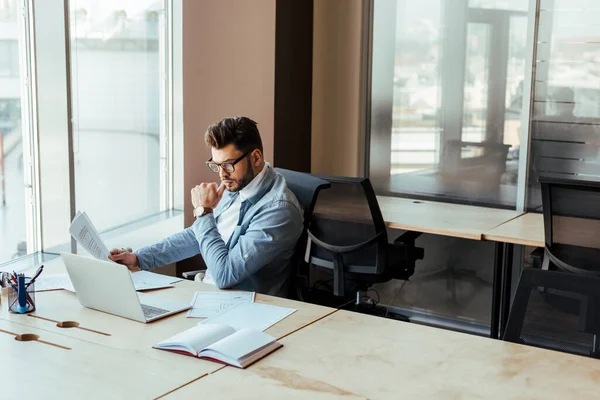 Vue en angle élevé du travailleur informatique concentré avec des papiers regardant un ordinateur portable à la table dans un espace de coworking — Photo de stock