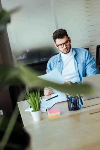 Focus selettivo del lavoratore IT concentrato in possesso di carte e guardando il laptop a tavola nello spazio di coworking — Foto stock