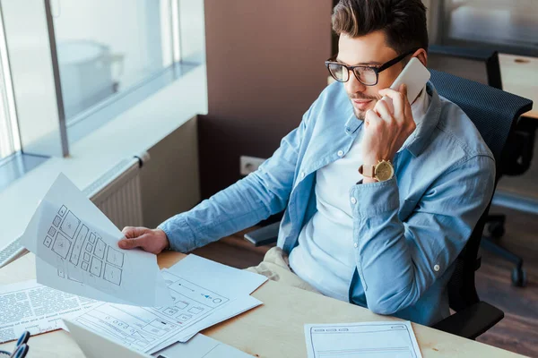 High angle view of IT worker holding papers, talking on smartphone and smiling at table in coworking space — Stock Photo