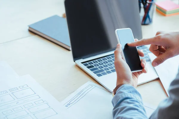 Vista ritagliata del lavoratore IT utilizzando smartphone a tavola con laptop e carte — Foto stock