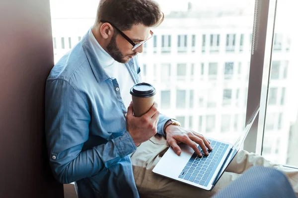 Selective focus of IT worker with paper cup of coffee working with laptop on on windowsill near windows — Stock Photo