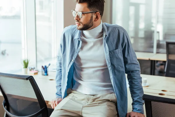 Thoughtful IT worker in glasses looking away and sitting on table in coworking space — Stock Photo