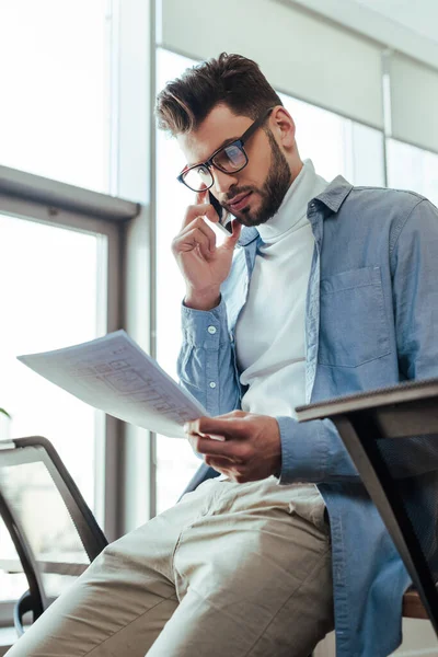 Low angle view of IT worker holding papers and talking on smartphone in coworking space — Stock Photo