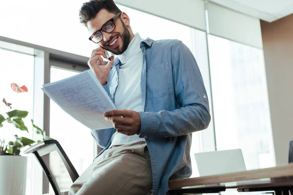 Low angle view of IT worker holding papers, talking on smartphone and smiling in coworking space — Stock Photo