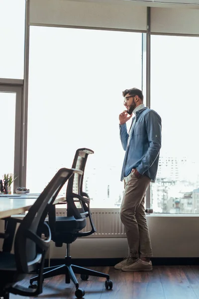 Trabajador de TI con la mano en el bolsillo hablando en el teléfono inteligente cerca de ventanas en el espacio de coworking - foto de stock