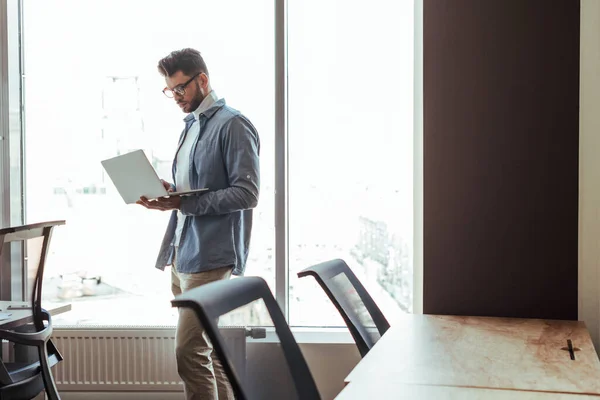 Trabajador de TI con portátil cerca de ventanas en espacio de coworking - foto de stock