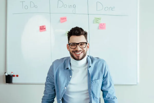 Scrum master looking at camera and smiling near white board with stickers and spreadsheet with to do, doing, done lettering — Stock Photo