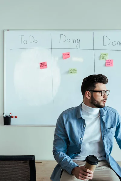 Scrum maître regardant loin avec tasse en papier de café près du tableau blanc avec feuille de calcul avec à faire, faire, lettrage fait et autocollants — Photo de stock