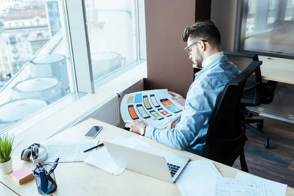 High angle view of UI designer with wove paper near table with laptop, smartphone and papers in coworking space — Stock Photo