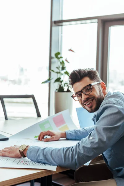 UI-Designer lächelt und blickt in die Kamera mit gewebten Papieren am Tisch — Stockfoto