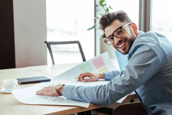 UI designer sorrindo e olhando para a câmera com papéis tecidos perto de xícara de café e notebook à mesa — Fotografia de Stock