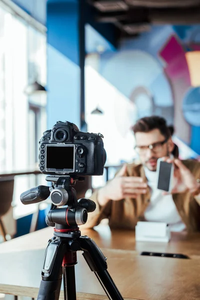 Selective focus of blogger in front of digital camera presenting smartphone in coworking space — Stock Photo