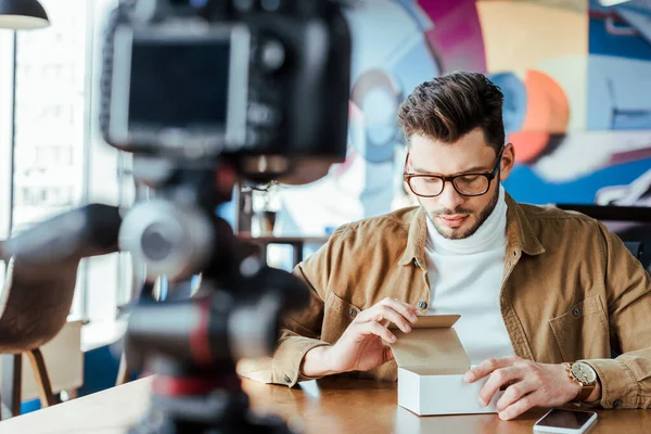 Selective focus of blogger looking at box near smartphone in front of digital camera at table in coworking space — Stock Photo