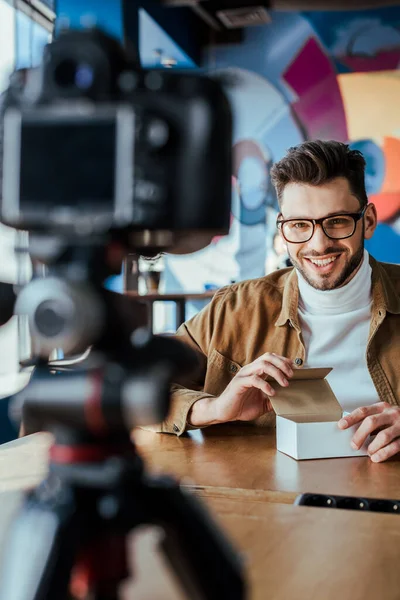 Selektiver Fokus des Bloggers mit geöffnetem Kasten lächelnd und Blick auf Digitalkamera am Tisch im Coworking Space — Stockfoto