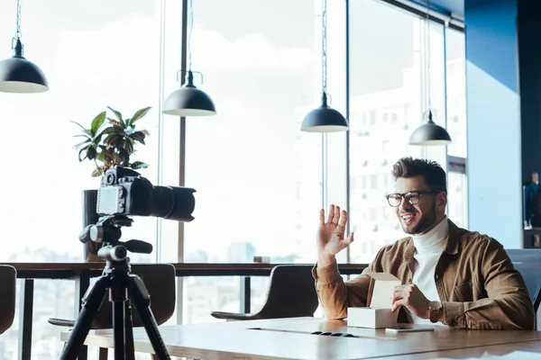 Blogger in front of digital camera waving hand, smiling and holding opened box at table in coworking space — Stock Photo