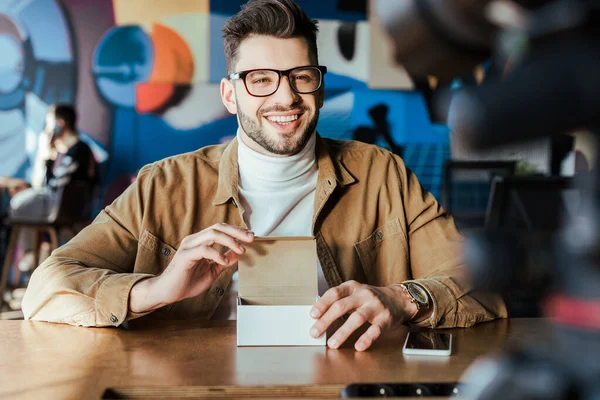 Selektiver Fokus eines Bloggers, der lächelt und eine geöffnete Box in der Nähe des Smartphones am Tisch im Coworking Space hält — Stockfoto