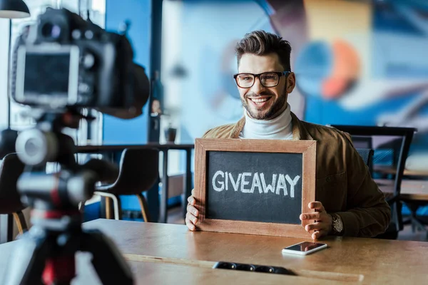 Selective focus of blogger showing board with giveaway lettering and smiling in front of  digital camera at table — Stock Photo