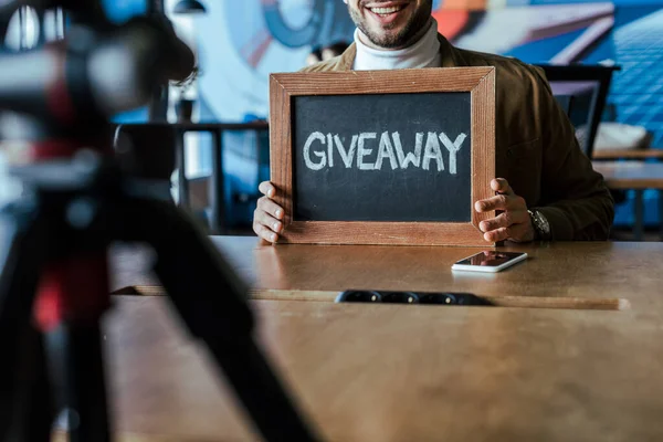 Cropped view of blogger showing board with giveaway lettering and smiling at table in coworking space — Stock Photo