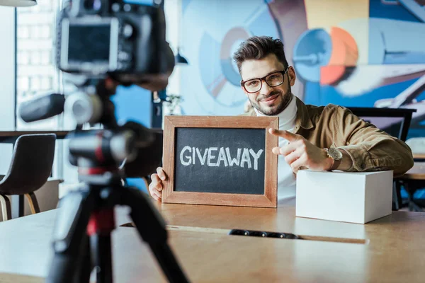 Selective focus of blogger showing board with giveaway lettering, smiling and pointing at digital camera at table — Stock Photo