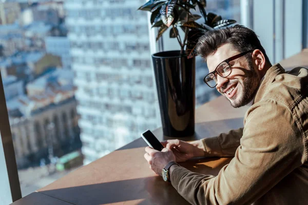 Visão de alto ângulo do trabalhador de TI com smartphone sorrindo à mesa com vaso perto da janela — Fotografia de Stock