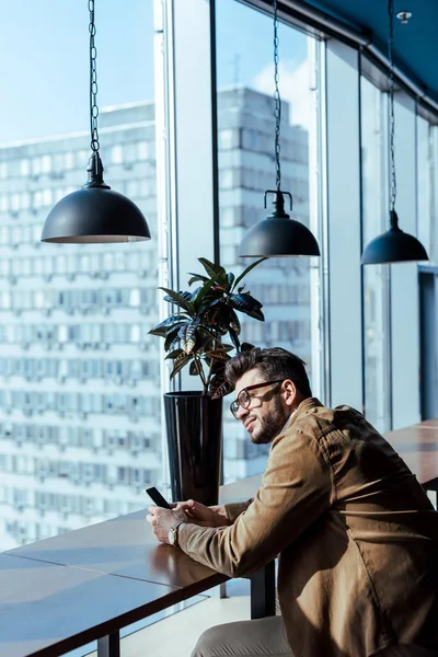 Nachdenklicher IT-Mitarbeiter mit Smartphone am Tisch in der Nähe von Fenstern im Coworking Space — Stockfoto